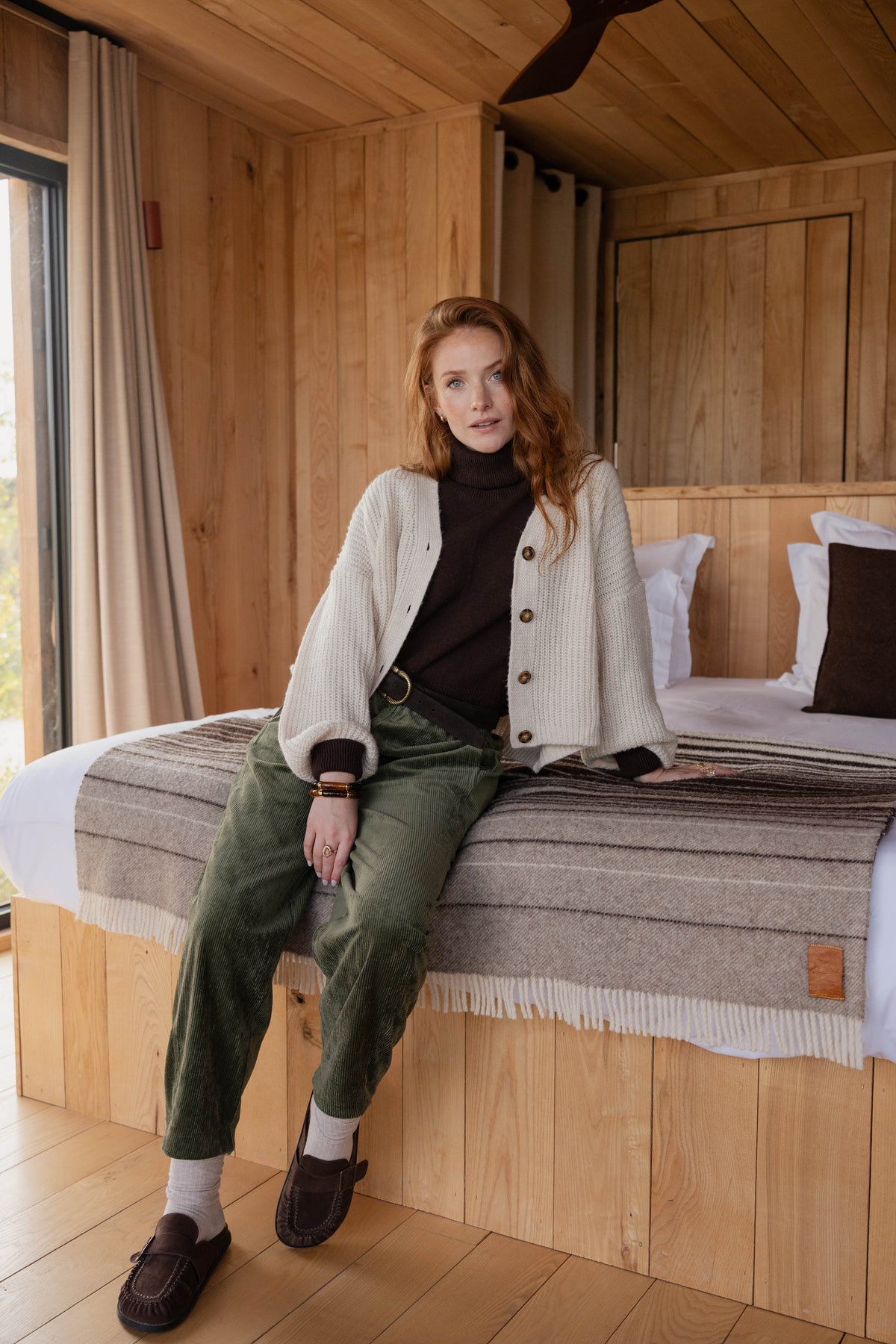 Gilet boutonné écru en maille avec un pull col roulé marron, une ceinture marron, un pantalon barrel kaki et une paire de chaussure marron 