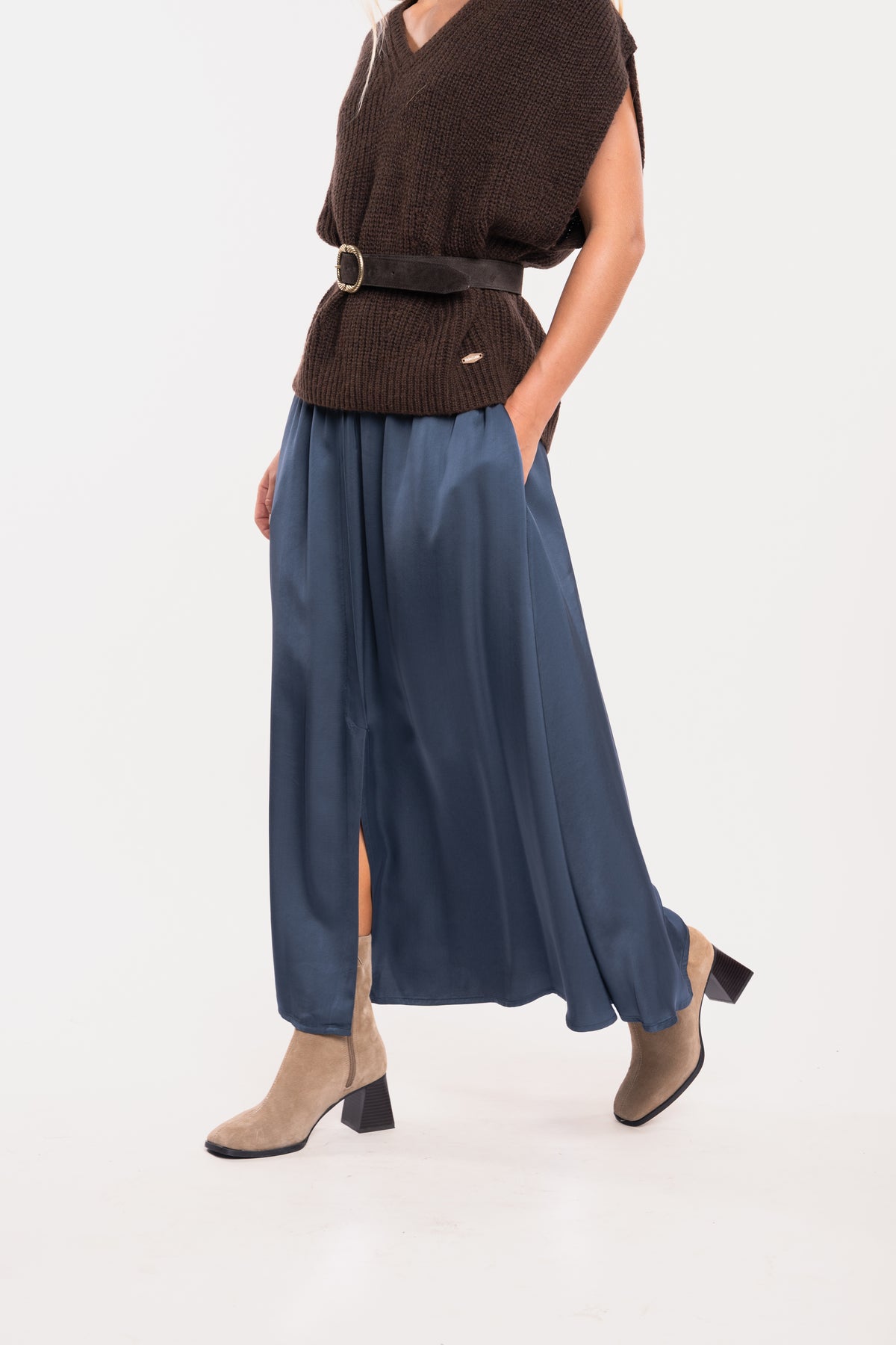 Un pull ample à col V marron, une ceinture en daim marron, une jupe midi fluide bleu nuit et une paire de bottes beiges 