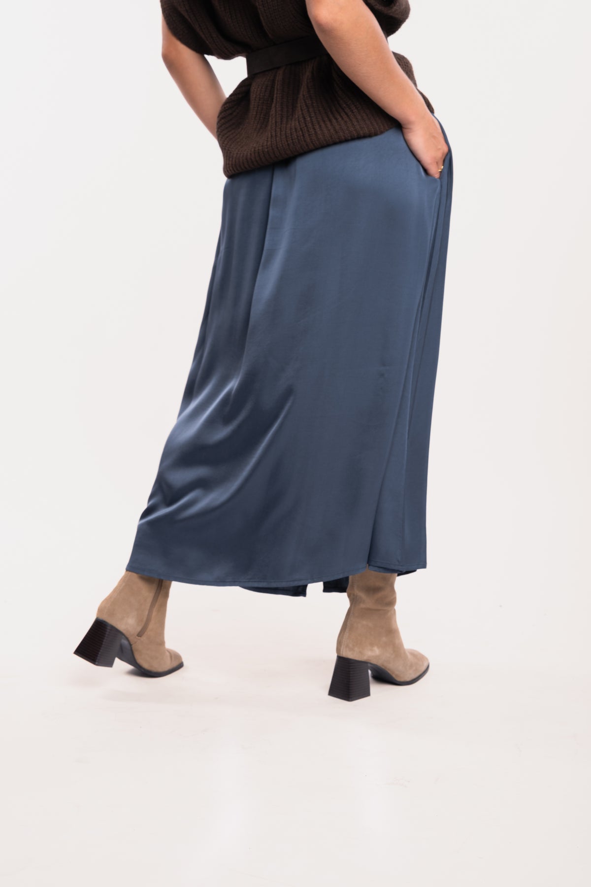 Un pull ample à col V marron, une ceinture en daim marron, une jupe midi fluide bleu nuit et une paire de bottes beiges de dos