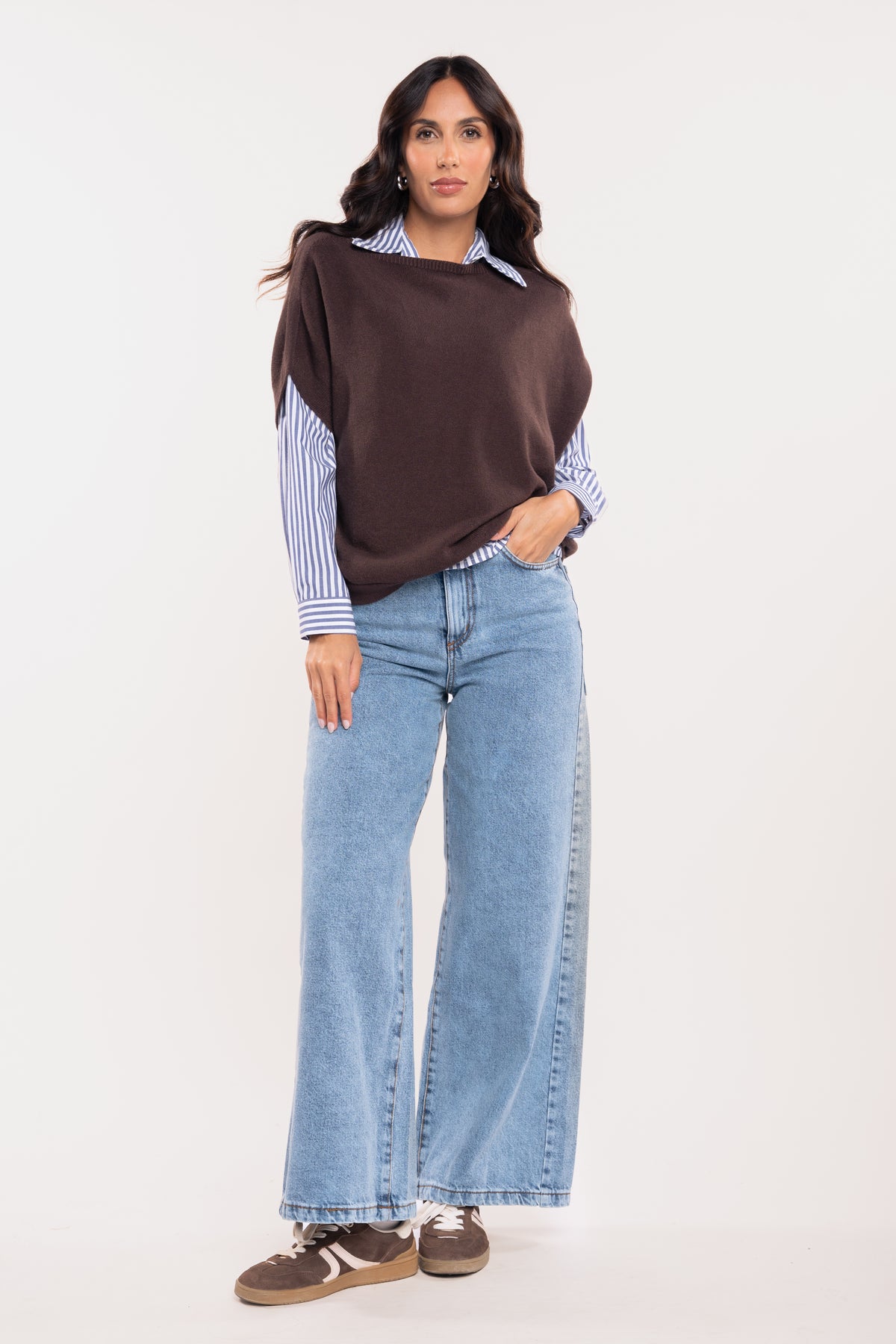 Pull sans manches marron avec chemise rayée bleu marine, jean évasé délavé clair et baskets marron 